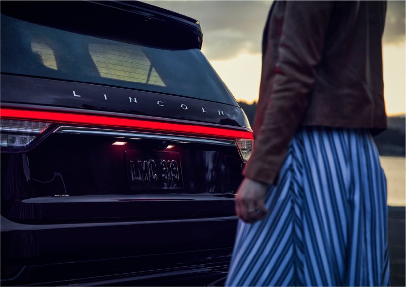 A person is shown near the rear of a 2023 Lincoln Aviator® SUV as the Lincoln Embrace illuminates the rear lights | Baldwin Lincoln in Covington LA