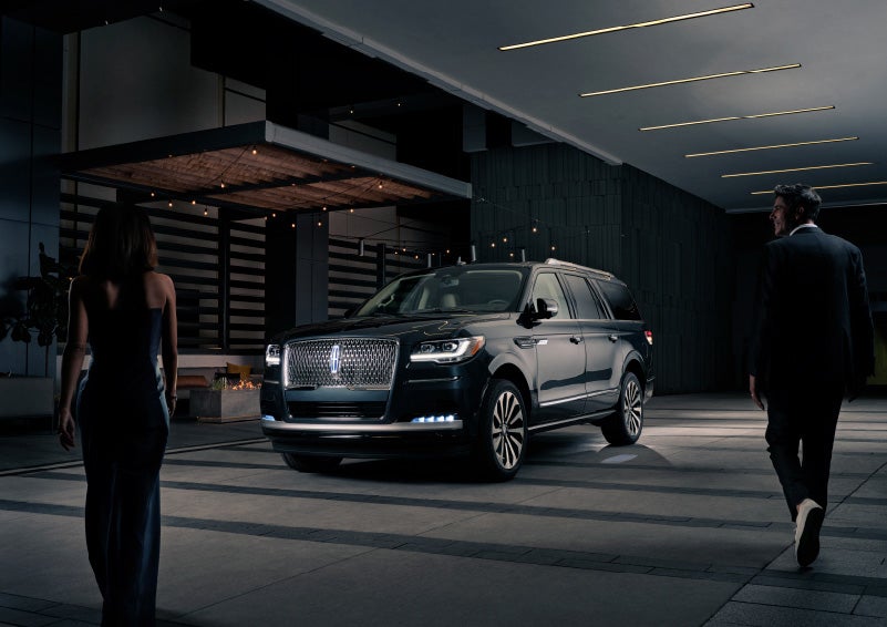 A man and woman in formal wear are approaching a 2022 Lincoln Navigator SUV at night as it lights with the Lincoln Embrace | Baldwin Lincoln in Covington LA