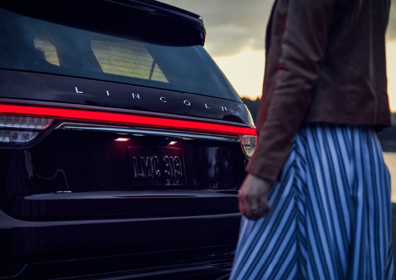 A person is shown near the rear of a 2024 Lincoln Aviator® SUV as the Lincoln Embrace illuminates the rear lights | Baldwin Lincoln in Covington LA